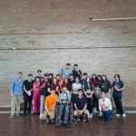 Grupo de personas ubicadas de frente, en su mayoría jóvenes, paradas y sentadas. Una de ellas en silla de ruedas.