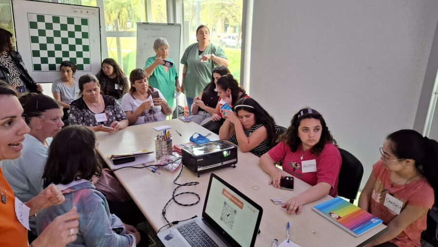 Fotografía de un taller presencial donde varias personas están sentadas alrededor de una mesa de trabajo con cuadernos, útiles y dispositivos móviles. En primer plano hay una computadora portátil conectada a un proyector, mostrando en pantalla el mensaje «HACEMOS UNA ENCUESTA» junto a un código QR y el logo de Por Igual +. Varias participantes llevan etiquetas con sus nombres y escuchan o participan activamente de la presentació