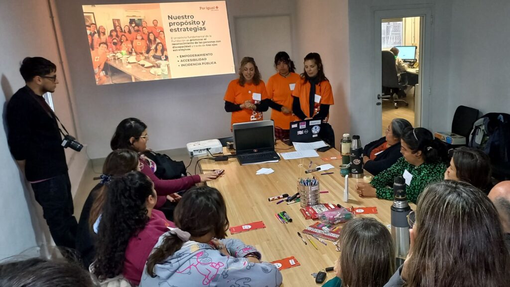 "Reunión de la Fundación Por Igual Más. Tres mujeres presentan los ejes estratégicos: Empoderamiento, Accesibilidad e Incidencia Pública, ante un equipo de trabajo en una mesa con computadoras y folletos."