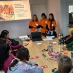 "Reunión de la Fundación Por Igual Más. Tres mujeres presentan los ejes estratégicos: Empoderamiento, Accesibilidad e Incidencia Pública, ante un equipo de trabajo en una mesa con computadoras y folletos."