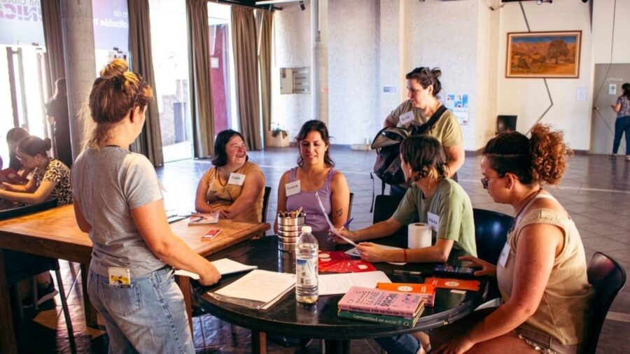 Un grupo de mujeres participa en un taller de mujeres, sentadas alrededor de una mesa redonda con papeles y libros en un salón amplio y luminoso."