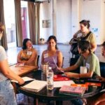 "Un grupo de mujeres participa en un taller de mujeres, sentadas alrededor de una mesa redonda con papeles y libros en un salón amplio y luminoso."