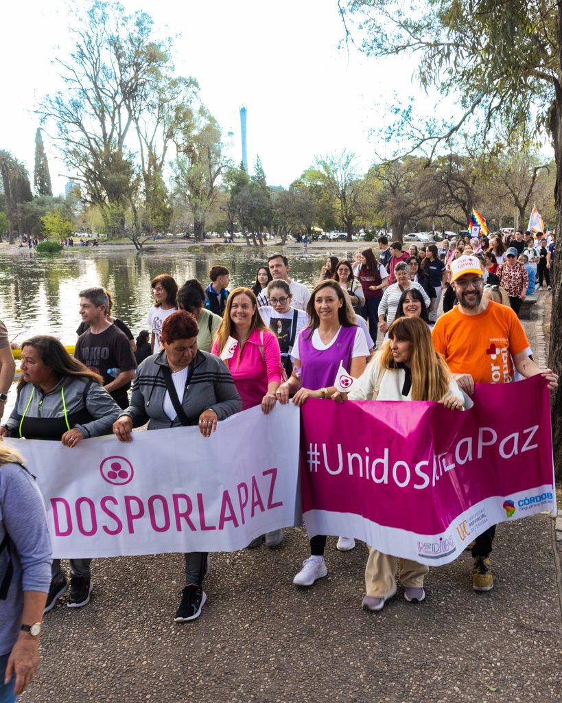Grupo de personas participando en una marcha al aire libre a la orilla de un lago en un parque. En primer plano, varias personas sostienen un par de pancartas; una blanca con la leyenda «DOSPORLAPAZ» y otra de color fucsia que dice «#UnidosPorLaPaz». Entre las personas que sostienen los carteles destaca un hombre con remera naranja y gorra de «Por Igual +