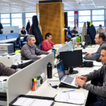 Vista amplia de una oficina de planta abierta iluminada con luz natural, donde varios empleados trabajan en escritorios individuales equipados con computadoras portátiles y monitores. En primer plano, se observa a varios hombres trabajando concentrados en sus estaciones de trabajo.